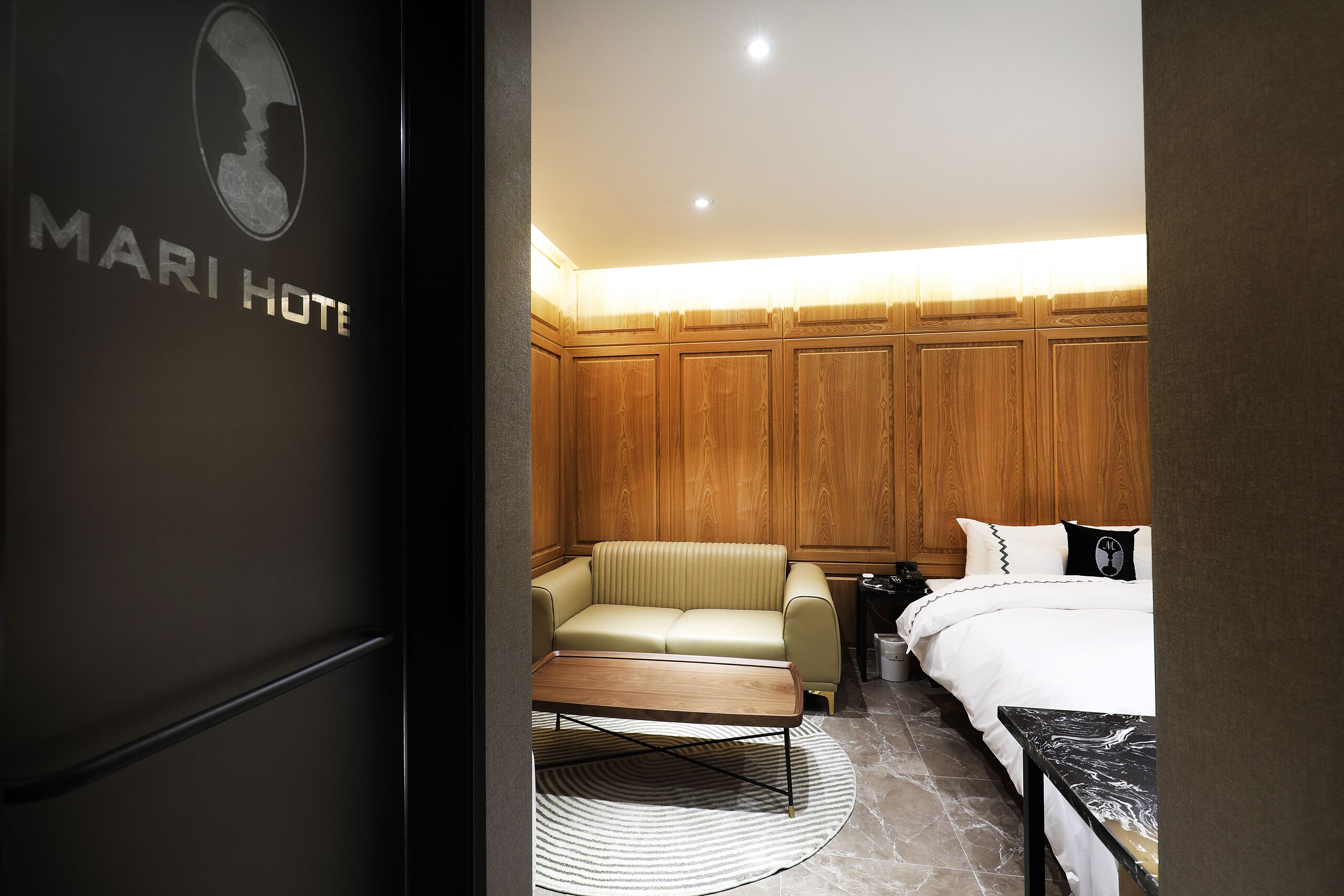 A hotel room with a wooden panel wall, a beige sofa, a bed with white bedding, and a marble coffee table.