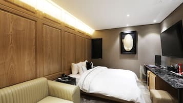 A hotel room with a bed, a sofa, a desk with a flat-screen TV, and a wall-mounted artwork.