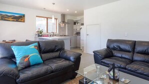 A modern living room with two black leather sofas, a glass coffee table, and a kitchen in the background.