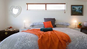 A bedroom with a bed, two bedside tables, a heart-shaped wall decoration, a window with blinds, and a painting on the wall.