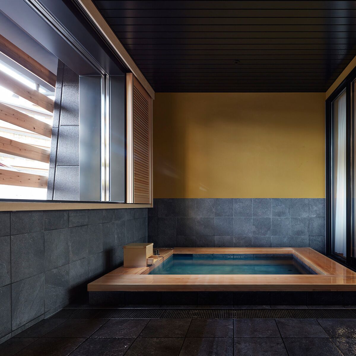 A modern bathroom with a wooden bathtub, dark tiled walls, and a window with blinds.