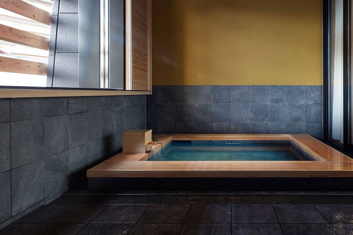 Ein modernes Badezimmer mit einer Holzbadewanne, dunkel gefliesten Wänden und einem Fenster mit Jalousien.