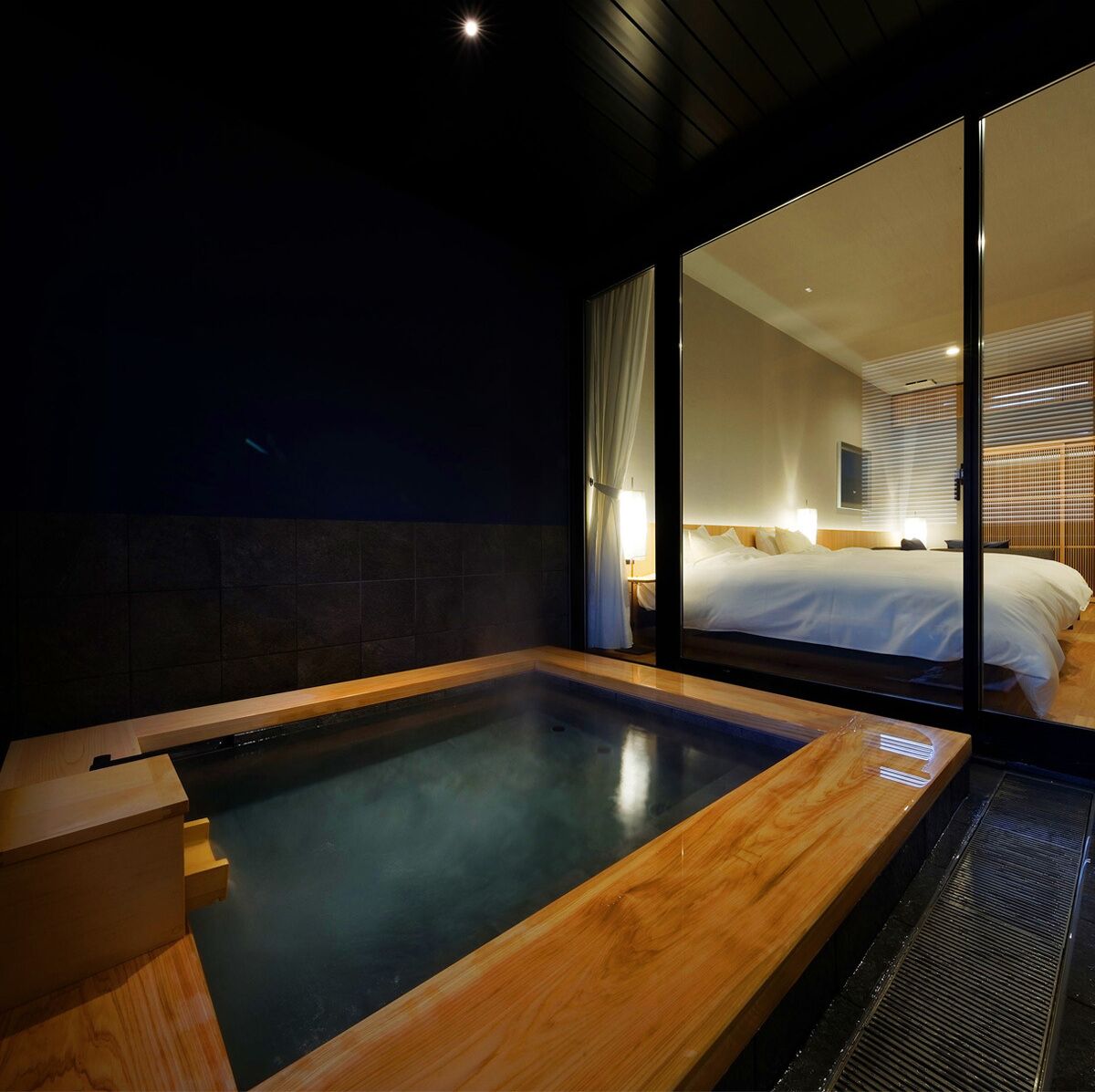 A modern hotel room with a large glass wall, a wooden bathtub, and a neatly made bed.