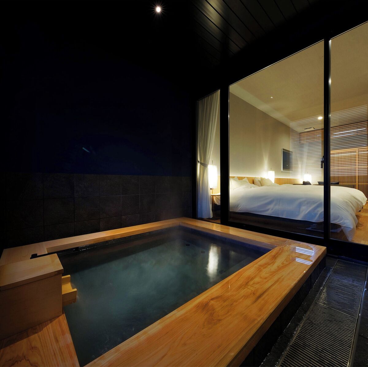 A modern hotel room with a large window, a wooden bathtub, and a bed with white linens.