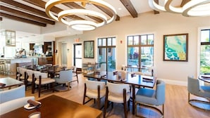 Moderni ruokailutila, jossa on useita pöytiä ja tuoleja, suuret ikkunat ja taustalla keittiö.