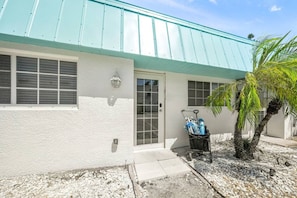 A building with a white exterior, a teal roof, a door, and a small garden area with a palm tree.