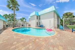 A swimming pool area with a curved pool edge, lounge chairs, and a building with a light-colored exterior and a blue roof. - Best location in SK Village, Walk to Beach (Sarasota)