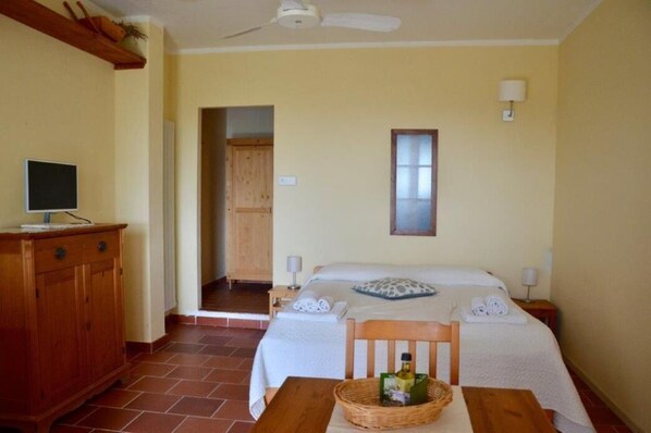 A hotel room with a bed, a wooden dresser, a television, and a table with a bottle and a basket.