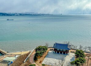 Un pavillon coréen traditionnel situé au bord de l’eau, offrant une vue sur les gratte-ciel d’une ville et un pont au loin.