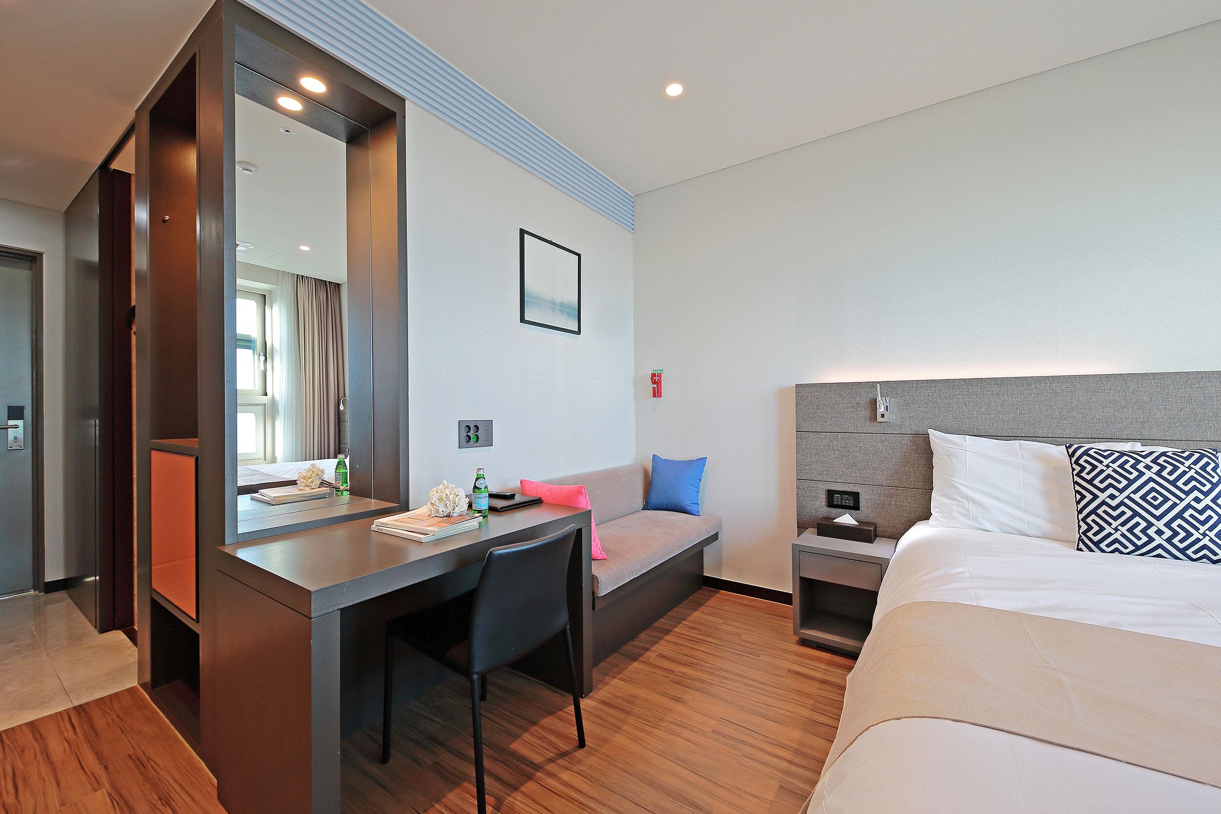A modern hotel room with a bed, a desk, a chair, a nightstand, and a large mirror.