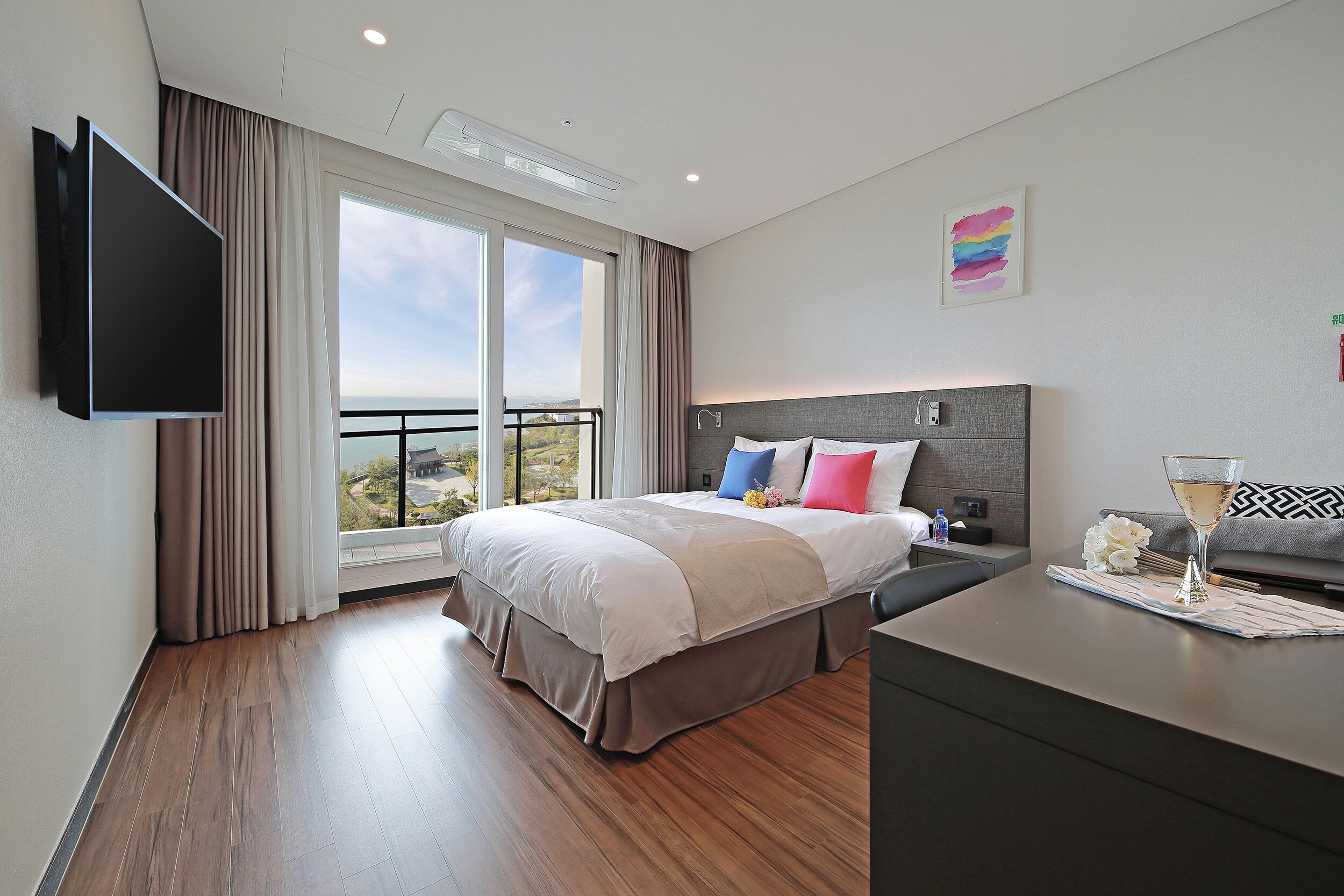 A modern hotel room with a large bed, a flat-screen TV, a balcony with a city view, and a minibar.