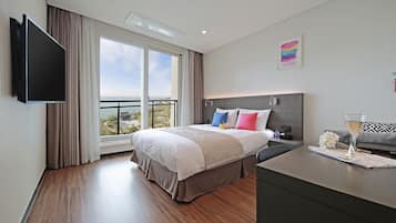 A modern hotel room with a large bed, a flat-screen TV, a balcony with a city view, and a minibar.