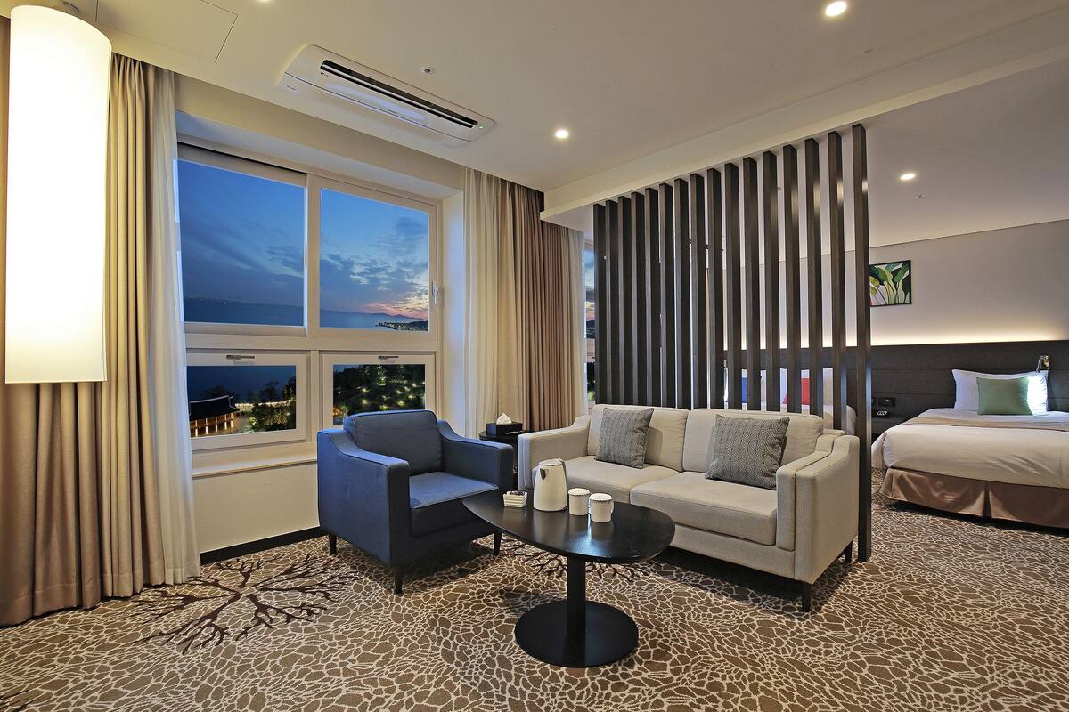 A hotel room with a sofa, armchair, and a small table with cups. There is a bed with a headboard and a window with curtains.