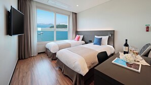Una habitación de hotel con dos camas, un televisor de pantalla plana, una mesa con una botella de vino y copas, y vista al mar.