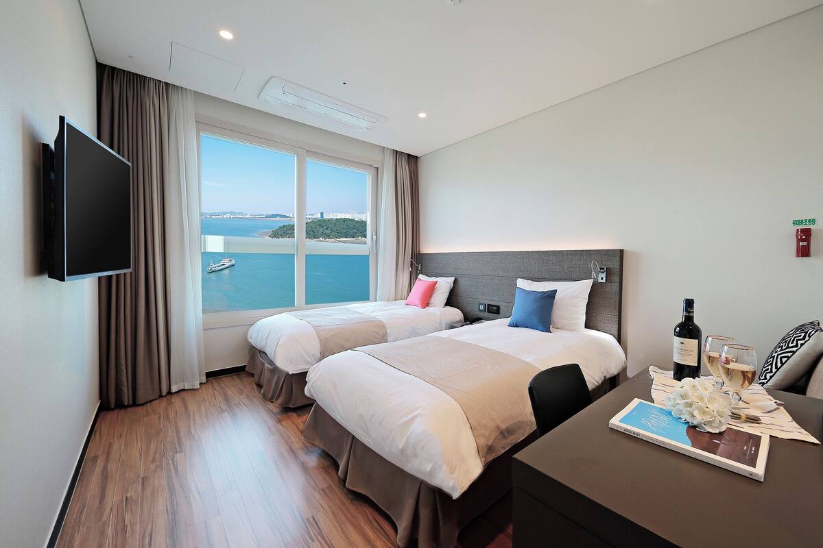 A hotel room with two beds, a flat-screen TV, a table with a bottle of wine and glasses, and a view of the sea.