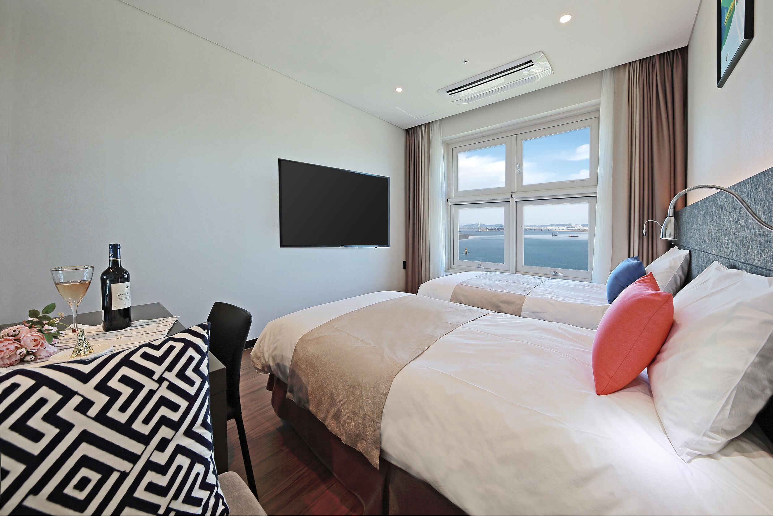 Une chambre d’hôtel avec deux lits, une télévision, une fenêtre donnant sur un beau paysage, et une table avec une bouteille et un verre.