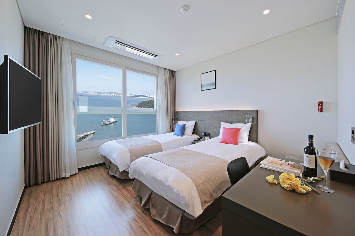 A hotel room with two beds, a large window with a view of the sea, a flat-screen TV, and a table with a bottle and glass.