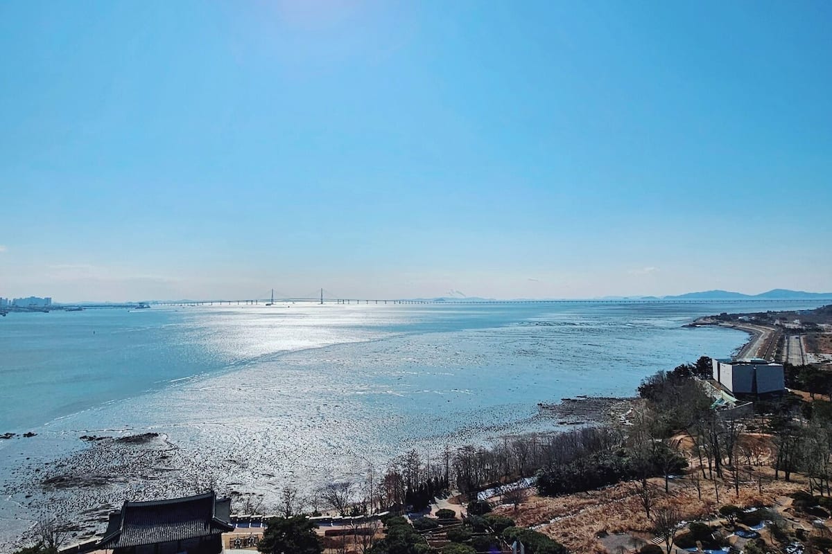 A coastal landscape with a bridge in the distance, a beach, and some buildings.