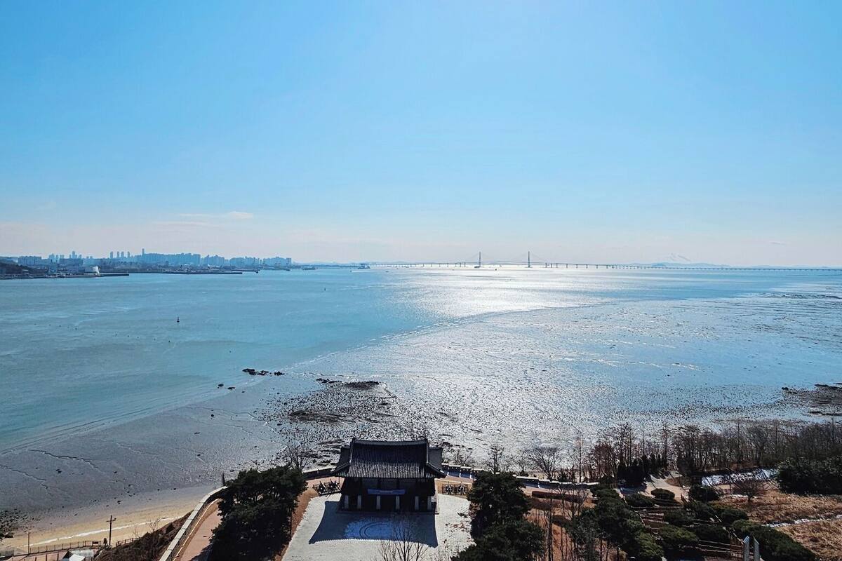 A coastal view with a traditional building in the foreground, a bridge in the distance, and a city skyline across the water.