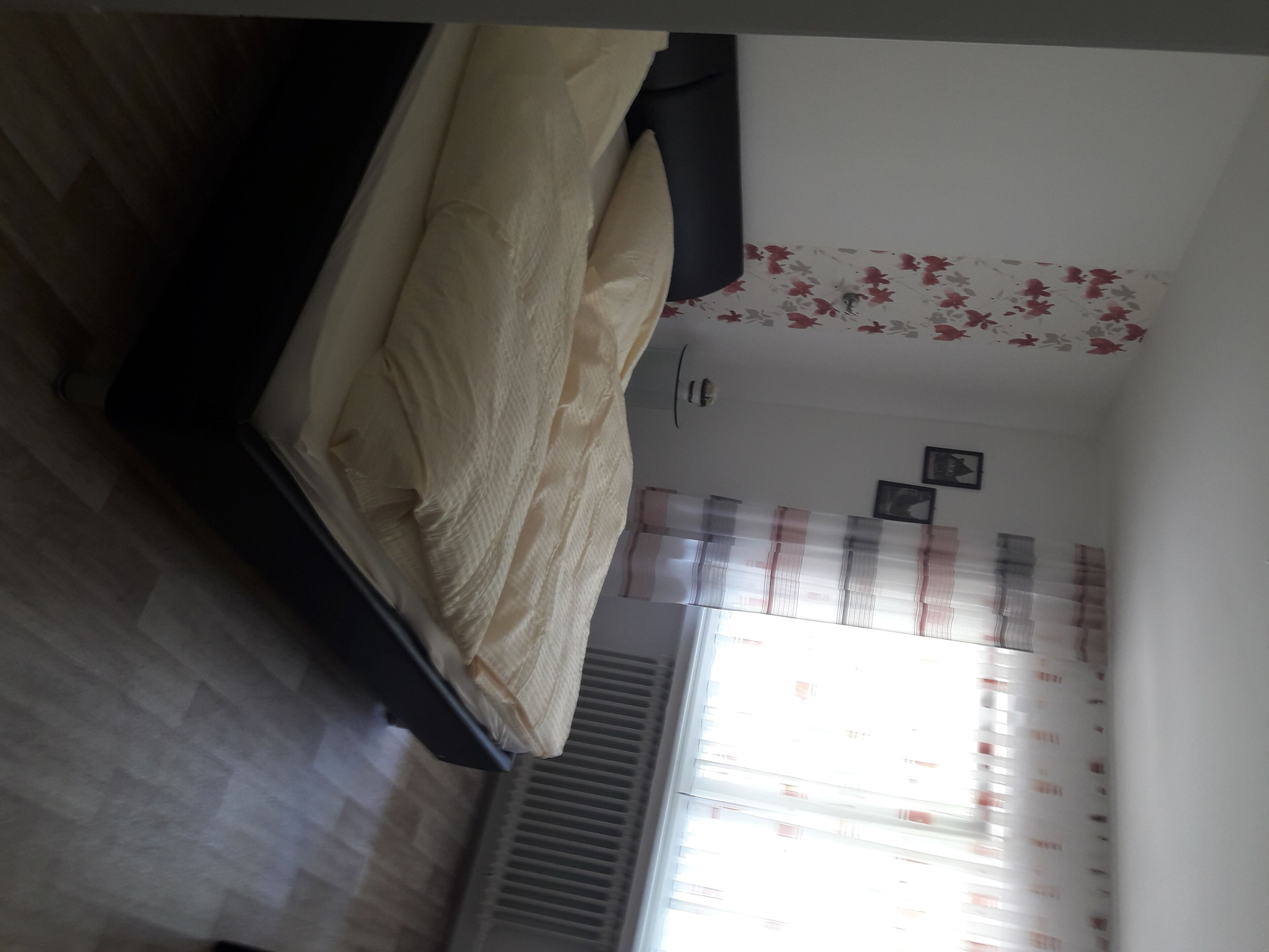 A bedroom with a bed, a radiator, a window with curtains, and a wall with a floral pattern.