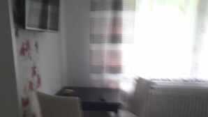 Quarto com sofá, poltrona, mesa pequena, TV fixada na parede e janela com cortinas.