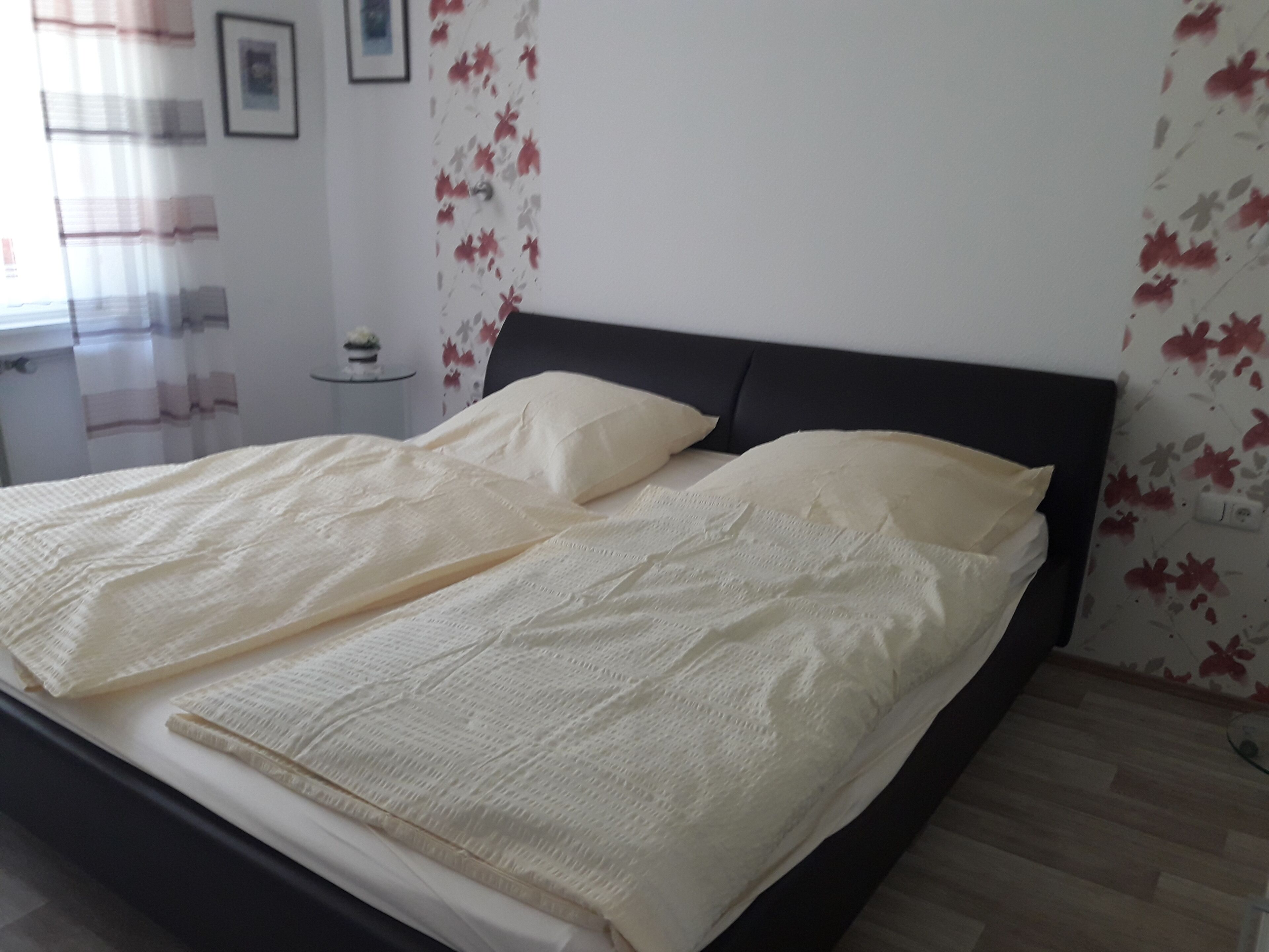 Una cama con edredón y almohadas blancos, una mesita de noche pequeña con lámpara, y papel tapiz floral en las paredes.