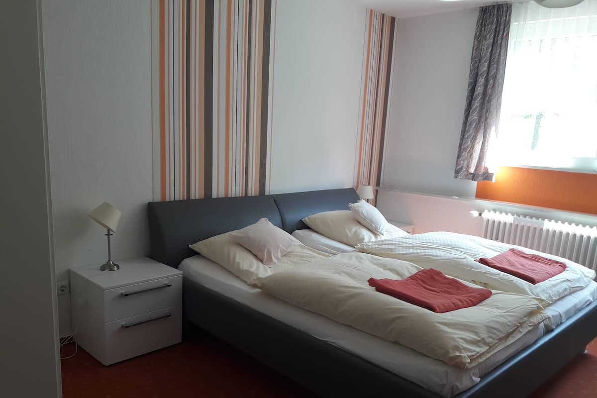 A neatly made bed with pillows and a blanket, a bedside table with a lamp, and a window with curtains.
