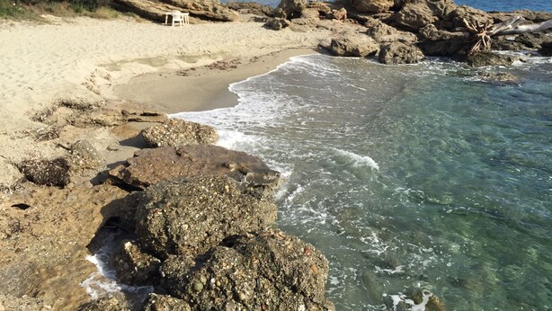 Ein Strand mit klarem Wasser, felsigem Ufer und einem weißen Stuhl im Sand.