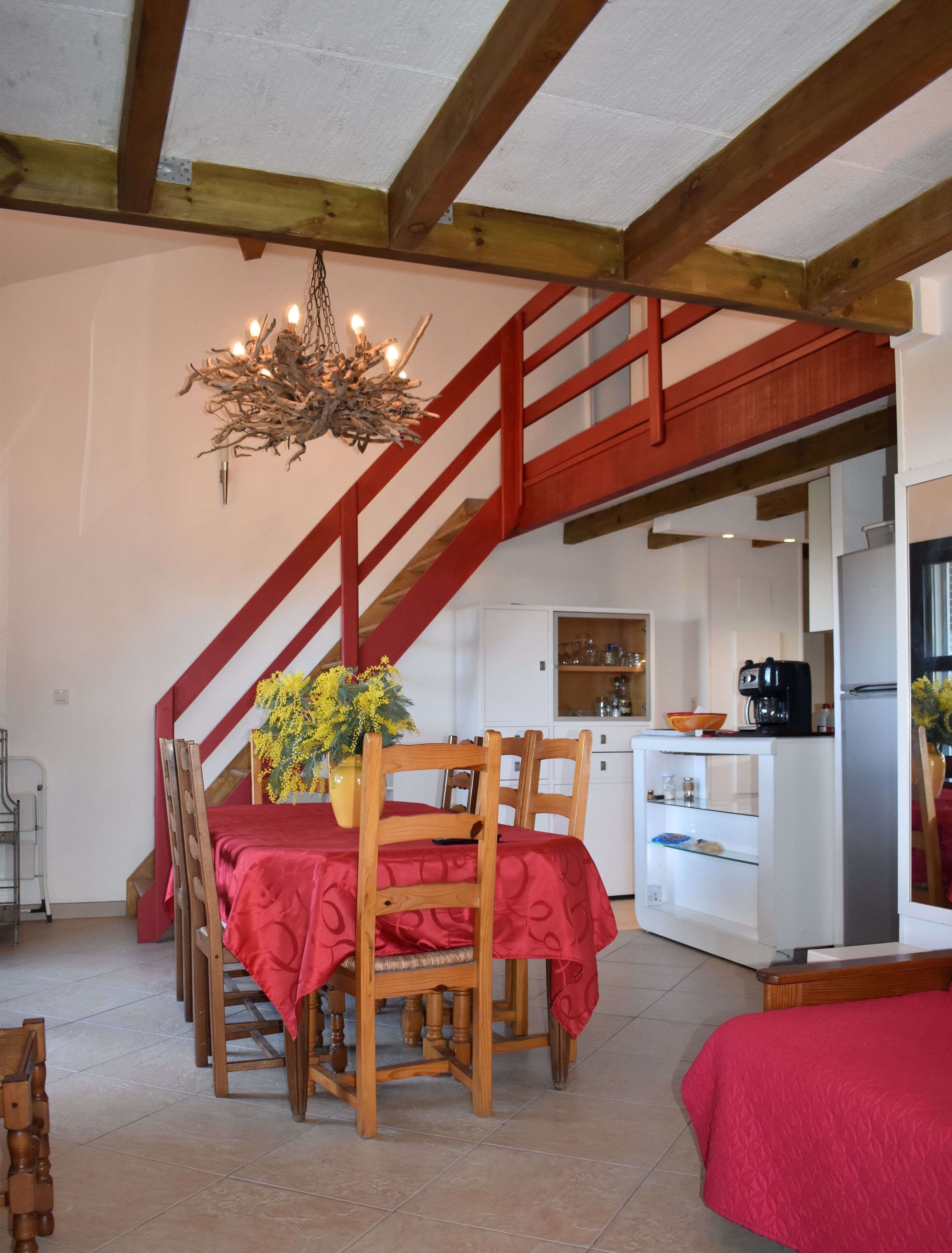 A dining area with a wooden table and chairs, a chandelier, and a staircase leading to a kitchen area.