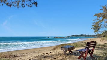 A beach with two wooden chairs and a small table, a clear blue sky, and calm sea waves.