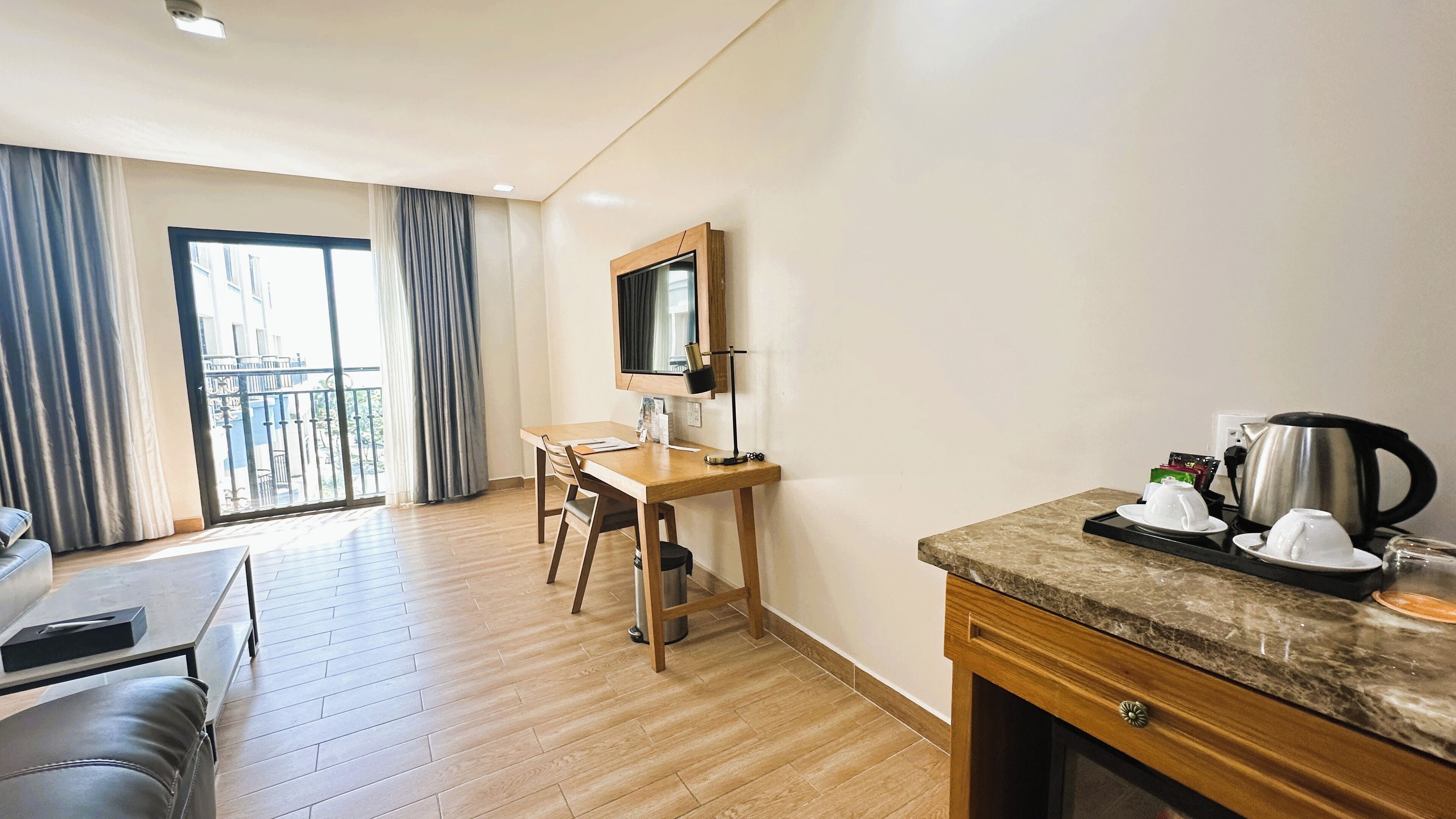 Habitación de hotel con escritorio de madera, una silla, una mesita con tetera y tazas, un sofá y un ventanal con cortinas.