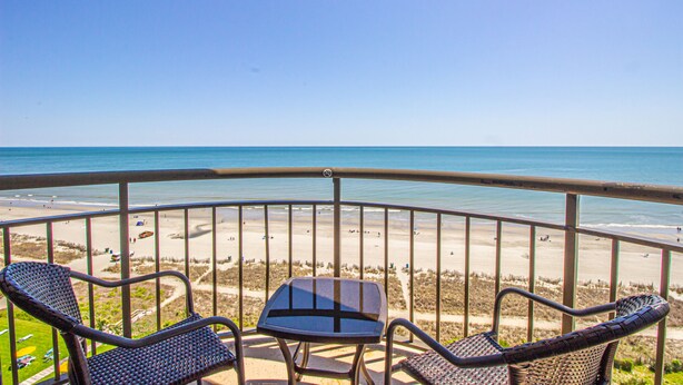 Ein Balkon mit zwei Korbstühlen und einem kleinen Tisch, mit Blick auf einen Strand mit Menschen und Sonnenschirmen.