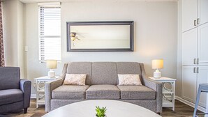 Ein Wohnzimmer mit grauem Sofa, zwei Beistelltischen mit Lampen, einem Couchtisch mit Pflanze und einem großen Spiegel im Bilderrahmen an der Wand.