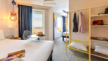 Habitación de hotel con cama, una mesita, una silla, una lámpara colgante, un armario y una ventana con cortinas.