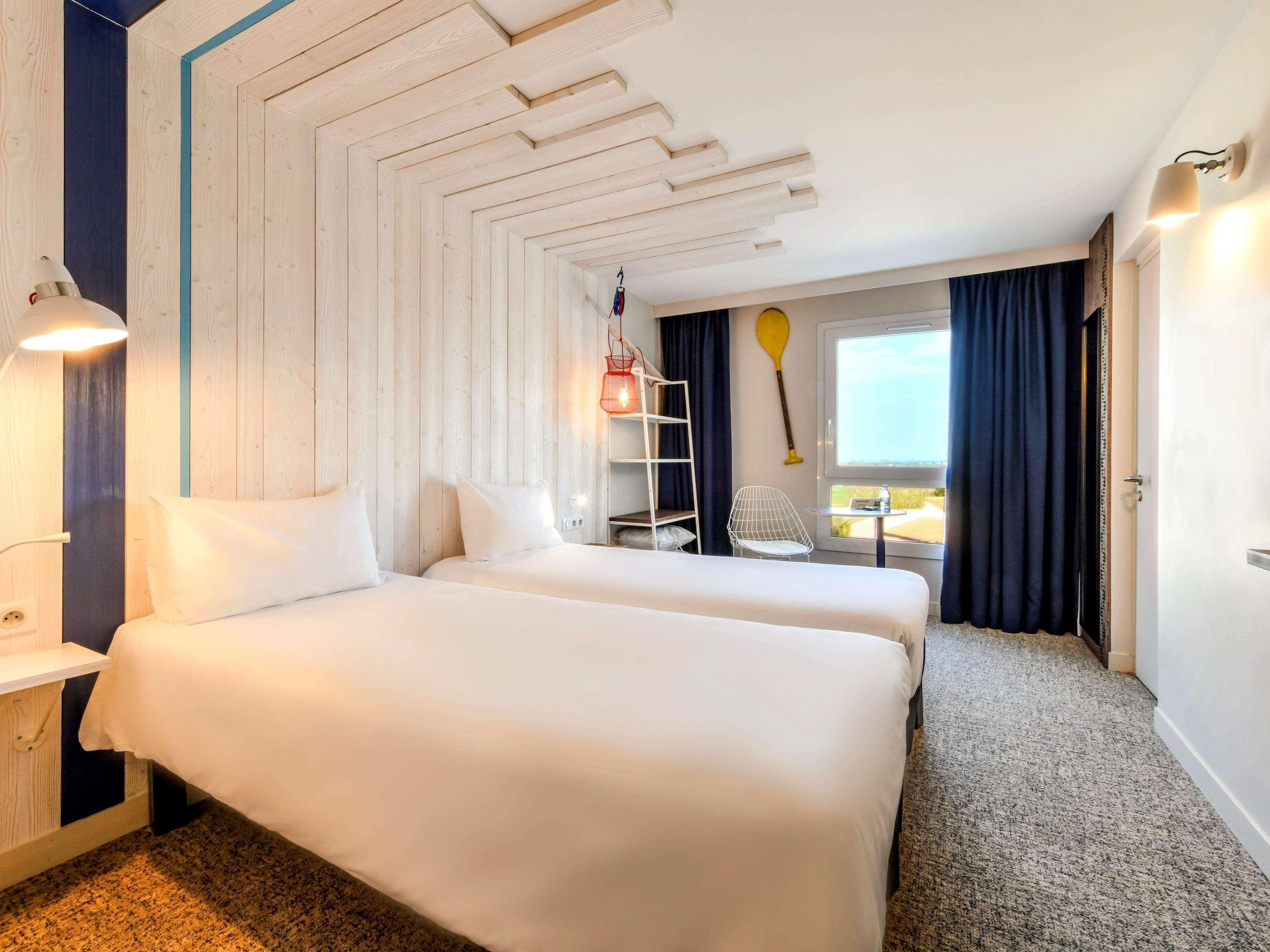 A hotel room with two beds, a desk, a chair, a window with curtains, a wall-mounted shelf, and a ceiling with a geometric design.