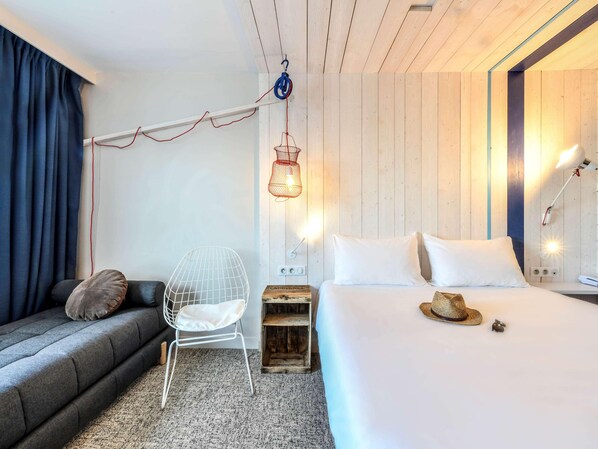 A modern hotel room with a bed, a chair, a hanging light fixture, and a wooden headboard. - Ibis Styles La Rochelle Thalasso Chatelaillon (Chatelaillon-Plage)