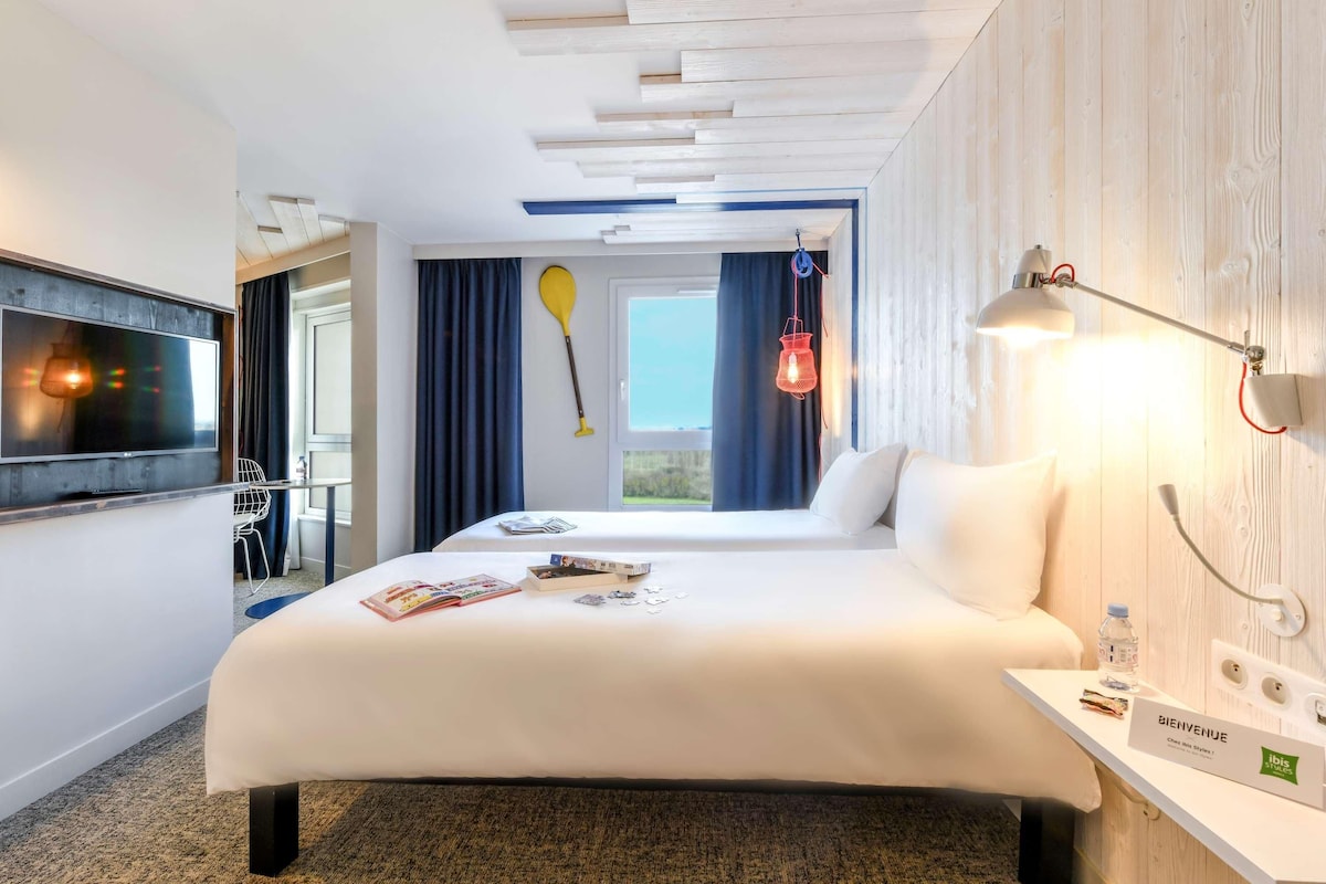 A hotel room with a bed, a desk, a TV, a window with curtains, a paddleboard, and a welcome sign.