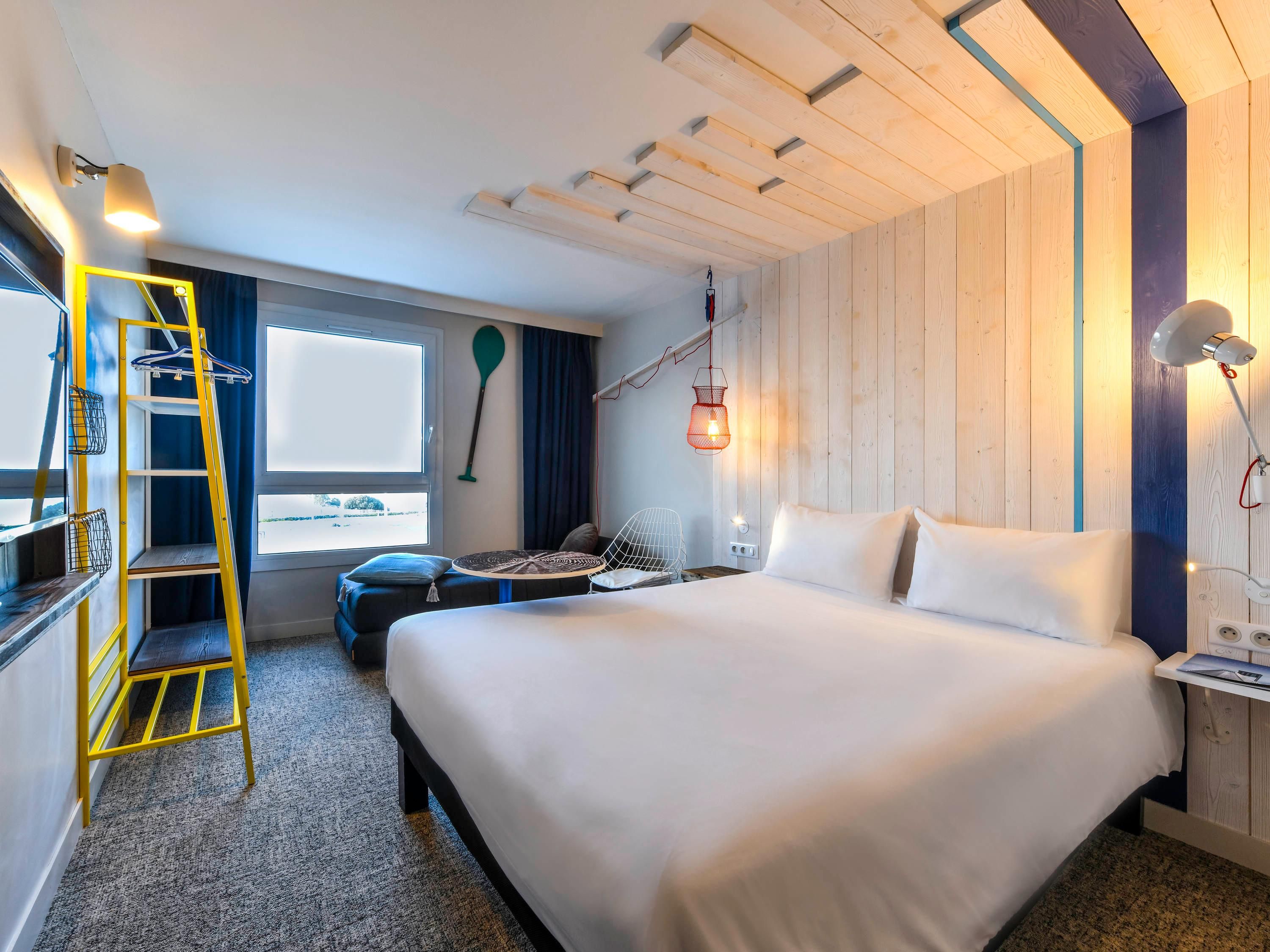 Habitació d'hotel amb un llit, una taula de treball, una cadira, una escala, una finestra i una làmpada penjada a la paret.