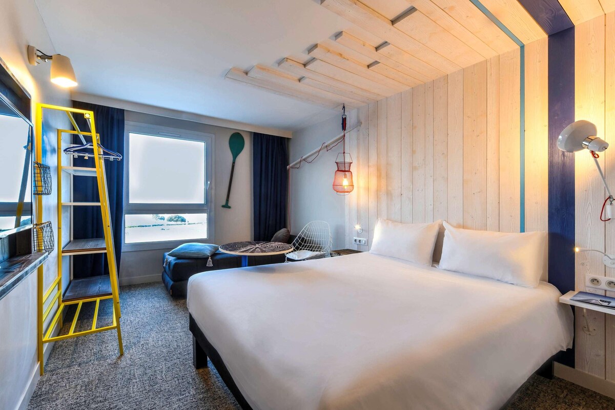 A hotel room with a bed, a desk with a chair, a small table, a ladder, a window, and a wall-mounted lamp.