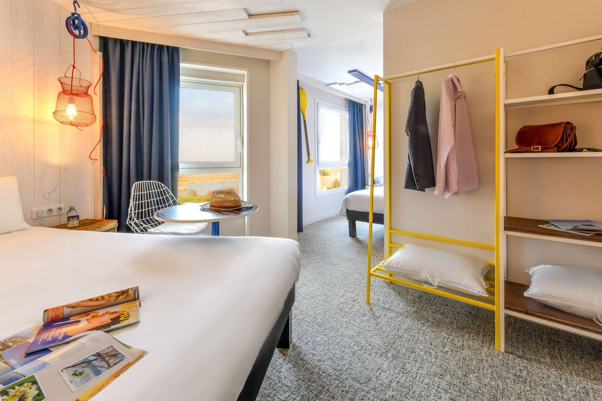 A hotel room with a bed, a small table, a chair, a hanging light, a wardrobe, and a window with curtains.