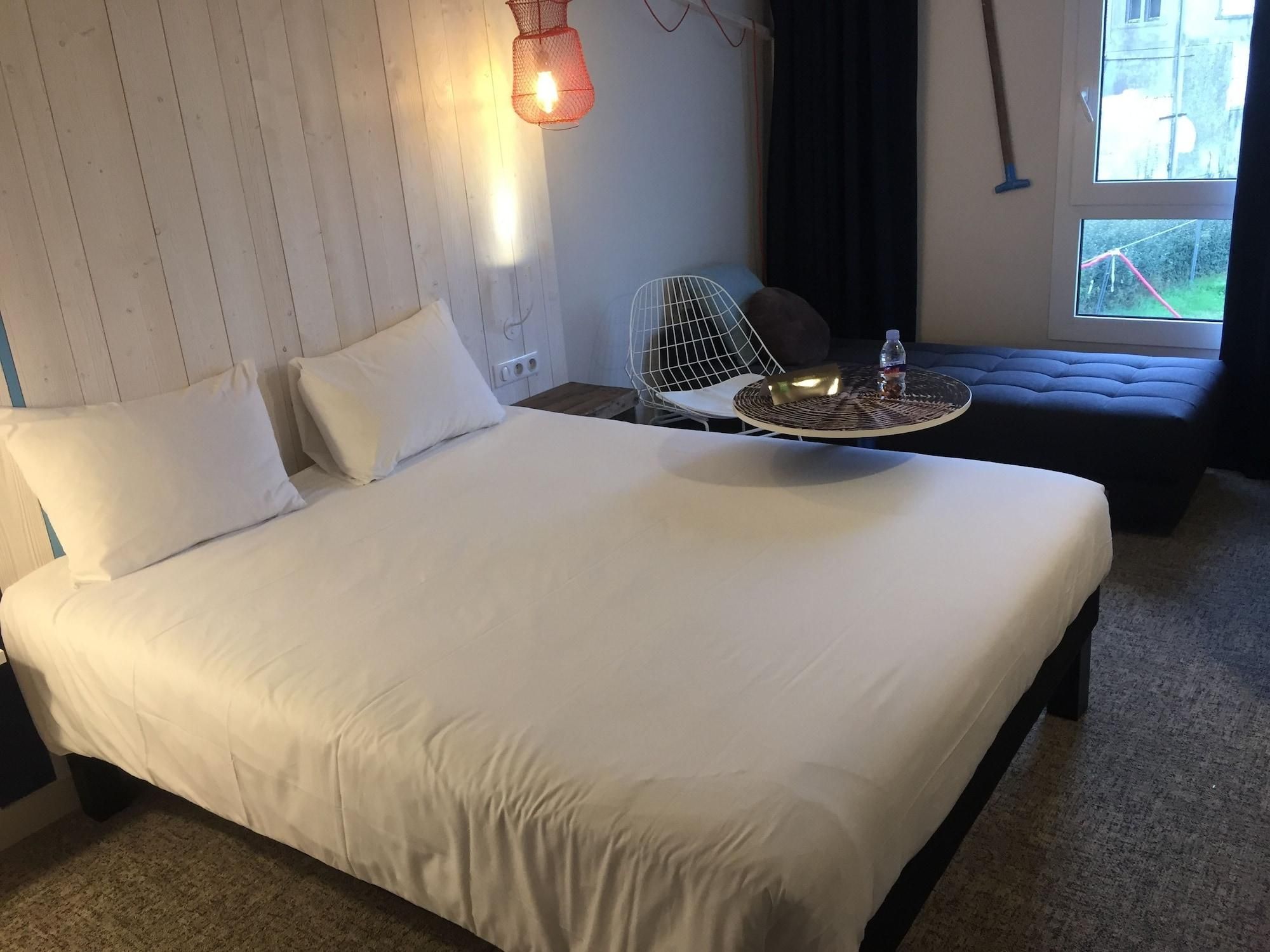 Ein Hotelzimmer mit einem großen Bett, einem kleinen runden Tisch mit einer Flasche, einem Stuhl und einem Fenster mit Blick nach draußen.