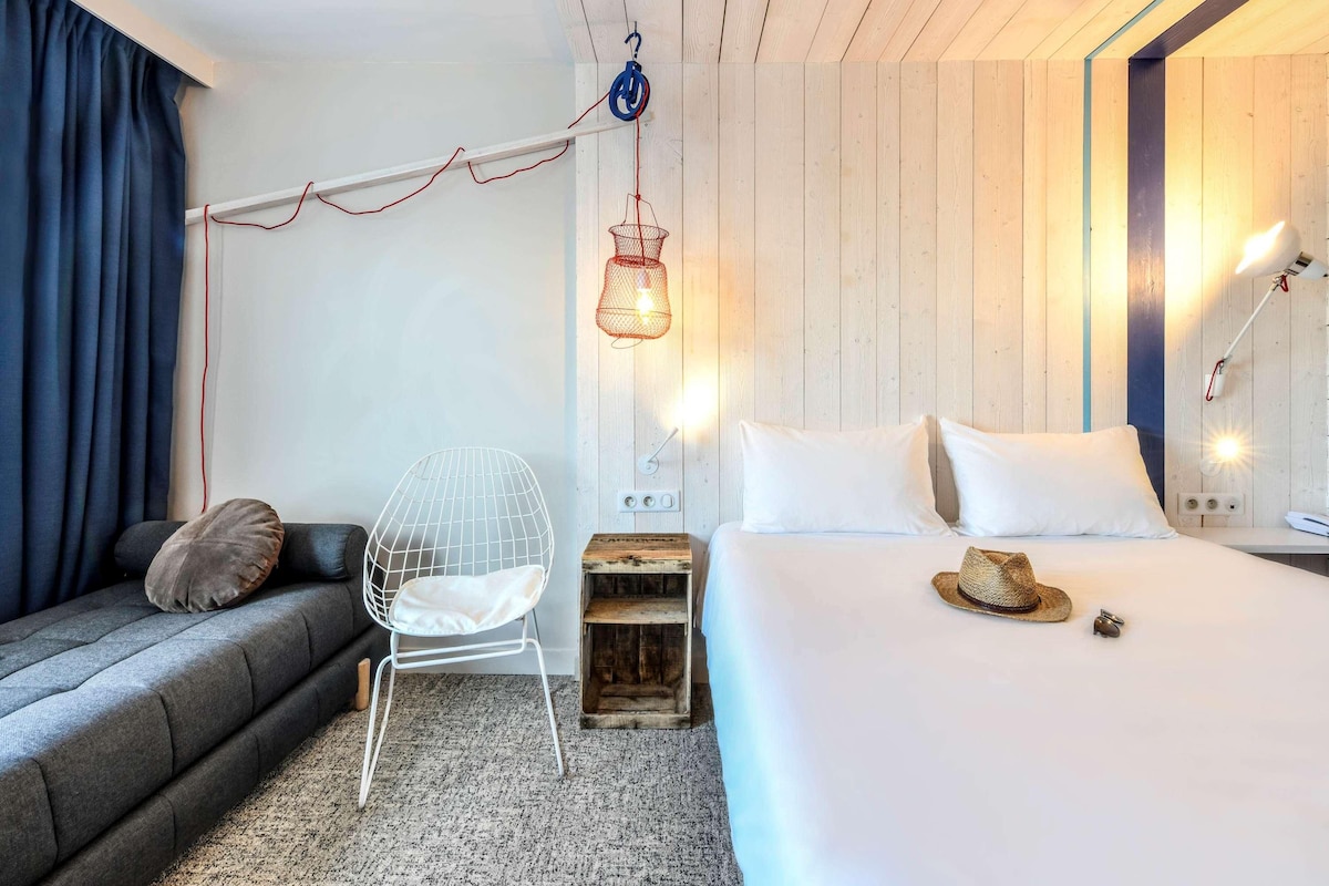 A modern hotel room with a bed, a chair, a hanging light fixture, and a wooden headboard.