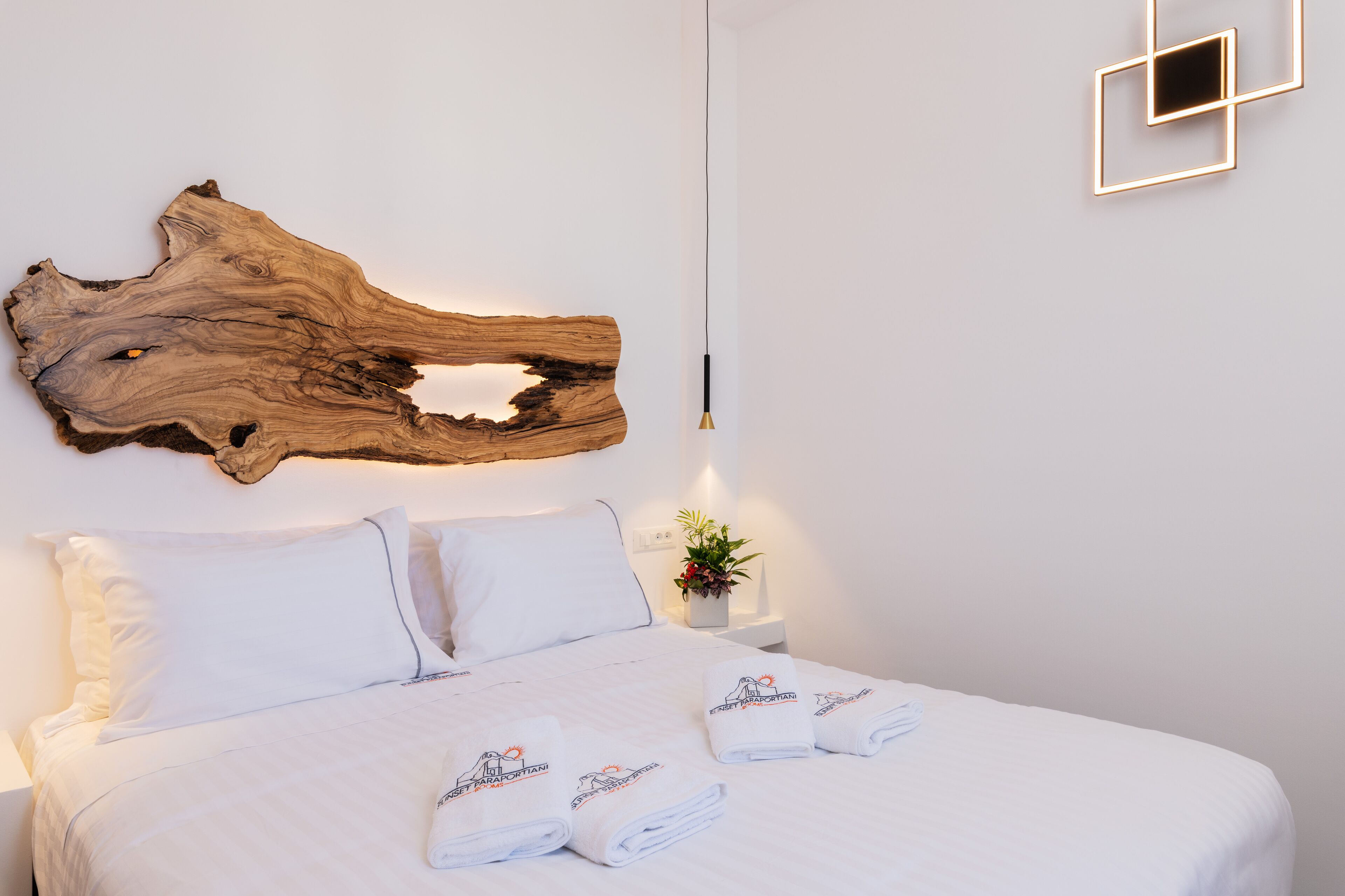 A neatly made bed with white linens and towels, a large piece of wood art on the wall, a bedside plant, and a modern light fixture.