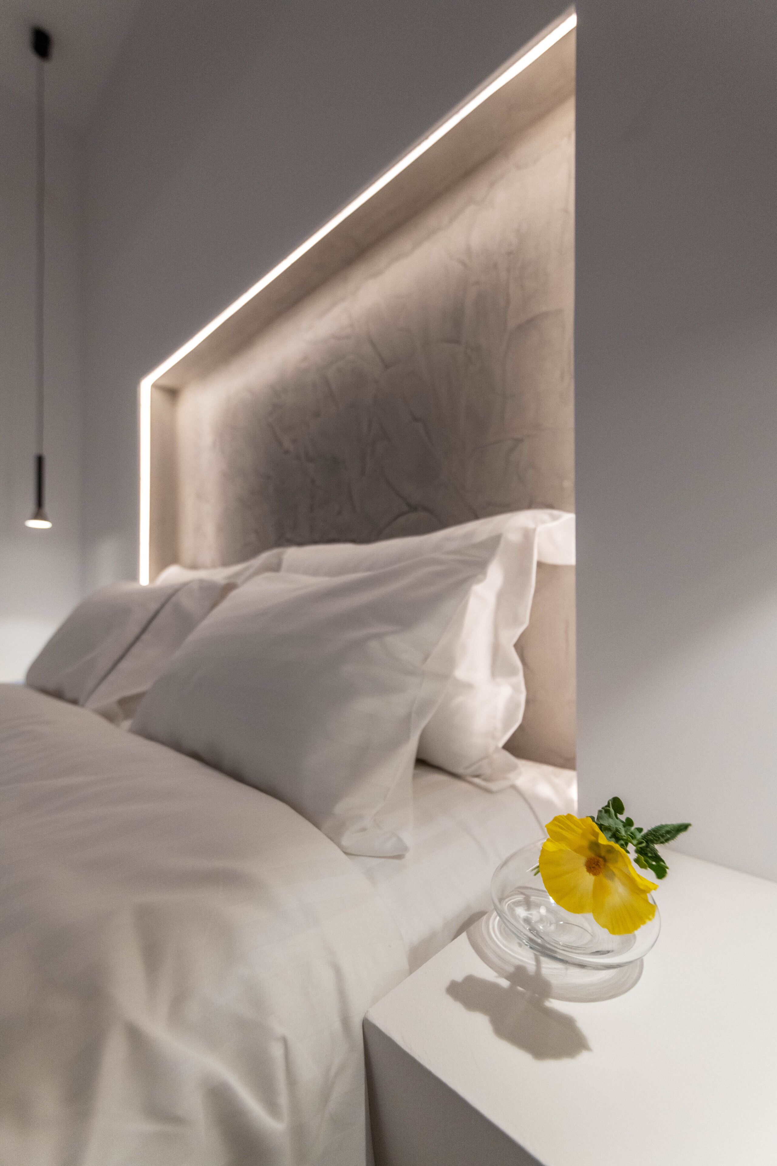 A modern bedroom with a minimalist design, featuring a bed with white linens, a clear glass vase with a yellow flower, and a geometric light fixture above the bed.
