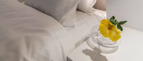 A modern bedroom with a minimalist design, featuring a bed with white linens, a clear glass vase with a yellow flower, and a geometric light fixture above the bed.