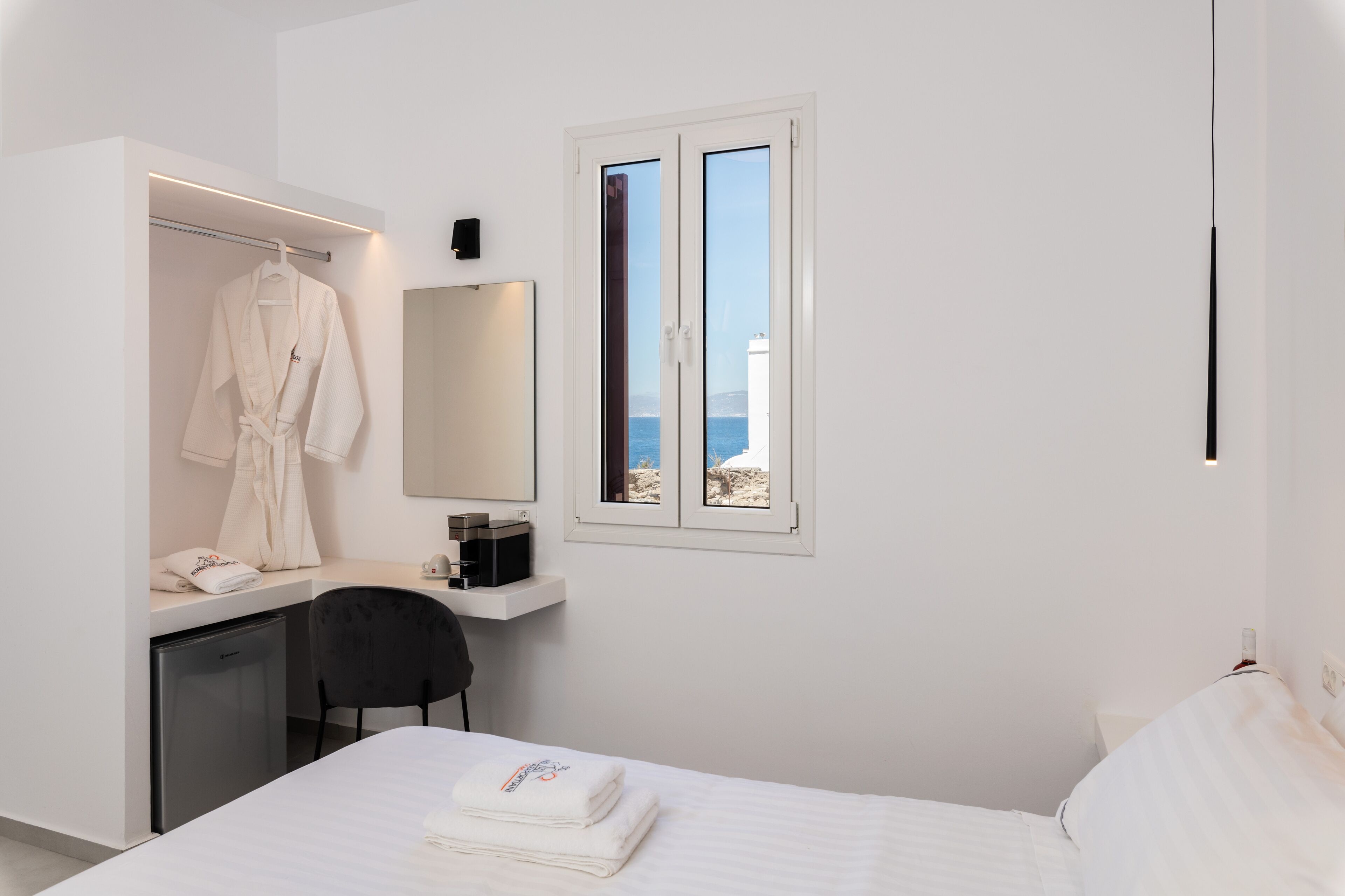 Une chambre d’hôtel avec un lit, un bureau et une chaise, une robe accrochée, un miroir et une fenêtre donnant sur la mer.