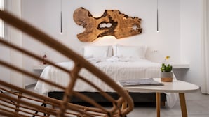 A modern bedroom with a large wooden wall art piece, a neatly made bed, a small table with a plant, and a wicker chair. - Sunset Paraportiani Rooms (Mykonos)