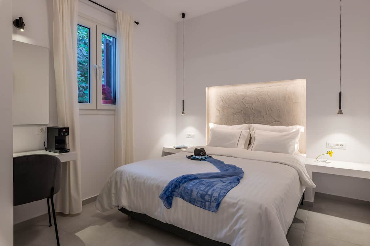 A modern hotel room with a large bed, a bedside table, a desk with a chair, a window with curtains, and a coffee machine.