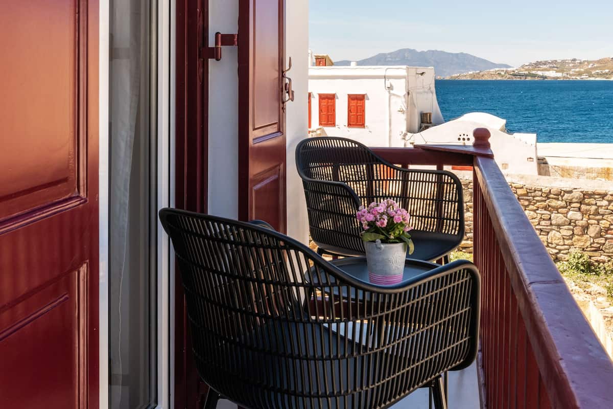 A balcony with two black chairs and a potted plant, overlooking a coastal view with buildings and the sea.