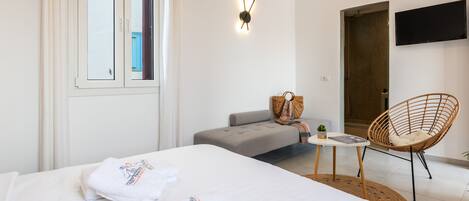 A modern hotel room with a large bed, a grey sofa, a wicker chair, a small table, and a TV mounted on the wall.